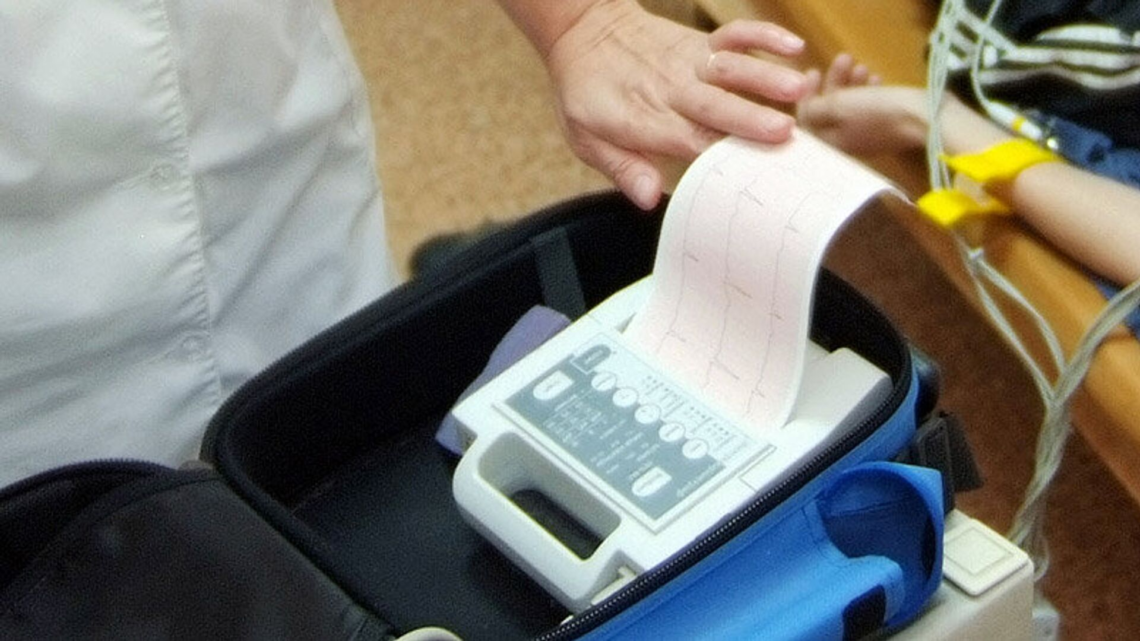 A medical professional holds an electrocardiogram (ECG) printout beside a patient with monitoring electrodes.